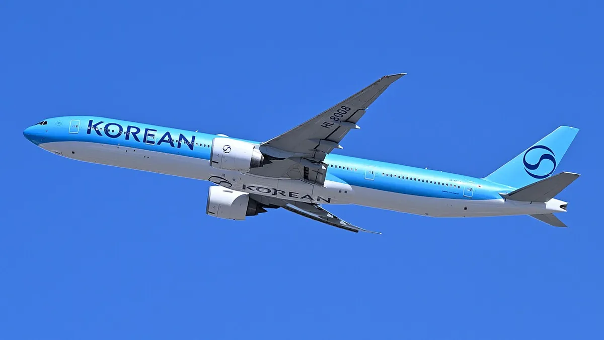 Korean Air Boeing 777-300ER climbing away from Boston Logan Airport in three-quarter view against a clear sky, showing its long stretched body and swept-to-a-point wingtips.