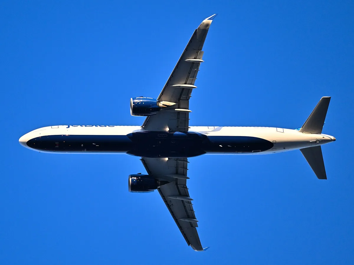 JetBlue Airbus A321-271NX seen directly from below against a clear blue sky, showing the underside outline, upturned wingtip blades, and oversized LEAP-1A engine pods.