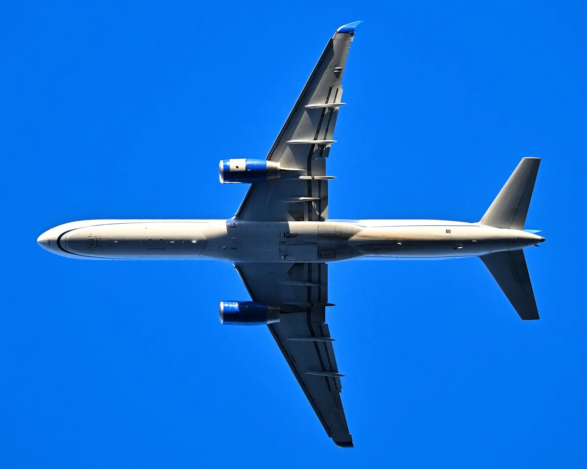 United Airlines Boeing 757-200 seen directly from below departing Boston, showing the underside outline and narrow, tapered wing.
