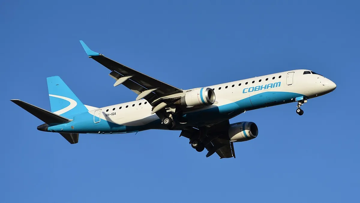 Cobham Aviation Embraer E190 seen from below-quarter on final approach, gear down, showing the underwing engines and upturned wingtip blades.