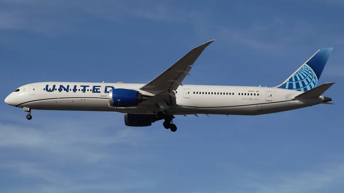United Airlines Boeing 787-10 N13013 on final approach with gear down, seen from a below-quarter angle against a clear sky, showing the underside of the wings and long body.