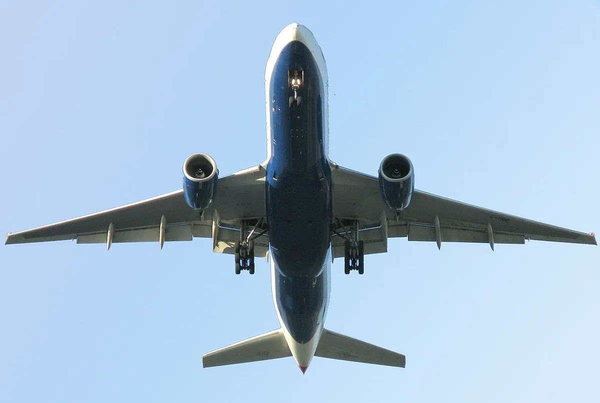 British Airways Boeing 777-200ER seen directly from below on final approach, showing the full underside outline, plain wingtips, and paired underwing engines.