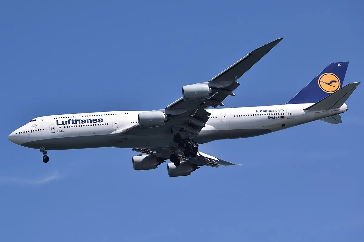 Lufthansa Boeing 747-8i seen from below on final approach, landing gear extended, showing the underside profile, saw-tooth-edged GEnx engine pods, and raked wingtips.