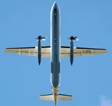 Flybe Dash 8 Q400 seen directly from below on takeoff, showing the high wing, twin propellers, and stretched body.