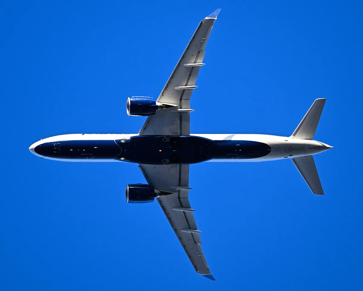 JetBlue Airbus A220-300 seen directly from below against a clear blue sky, showing the underside outline, oversized engine pods, and curved wingtip blades.